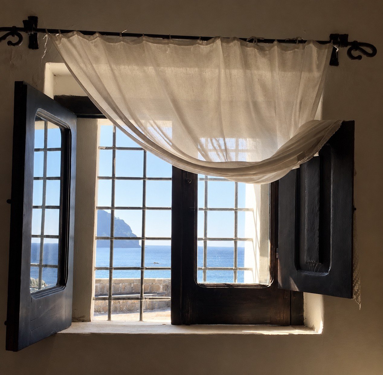An open wooden window with a linen curtain and a view of the sea