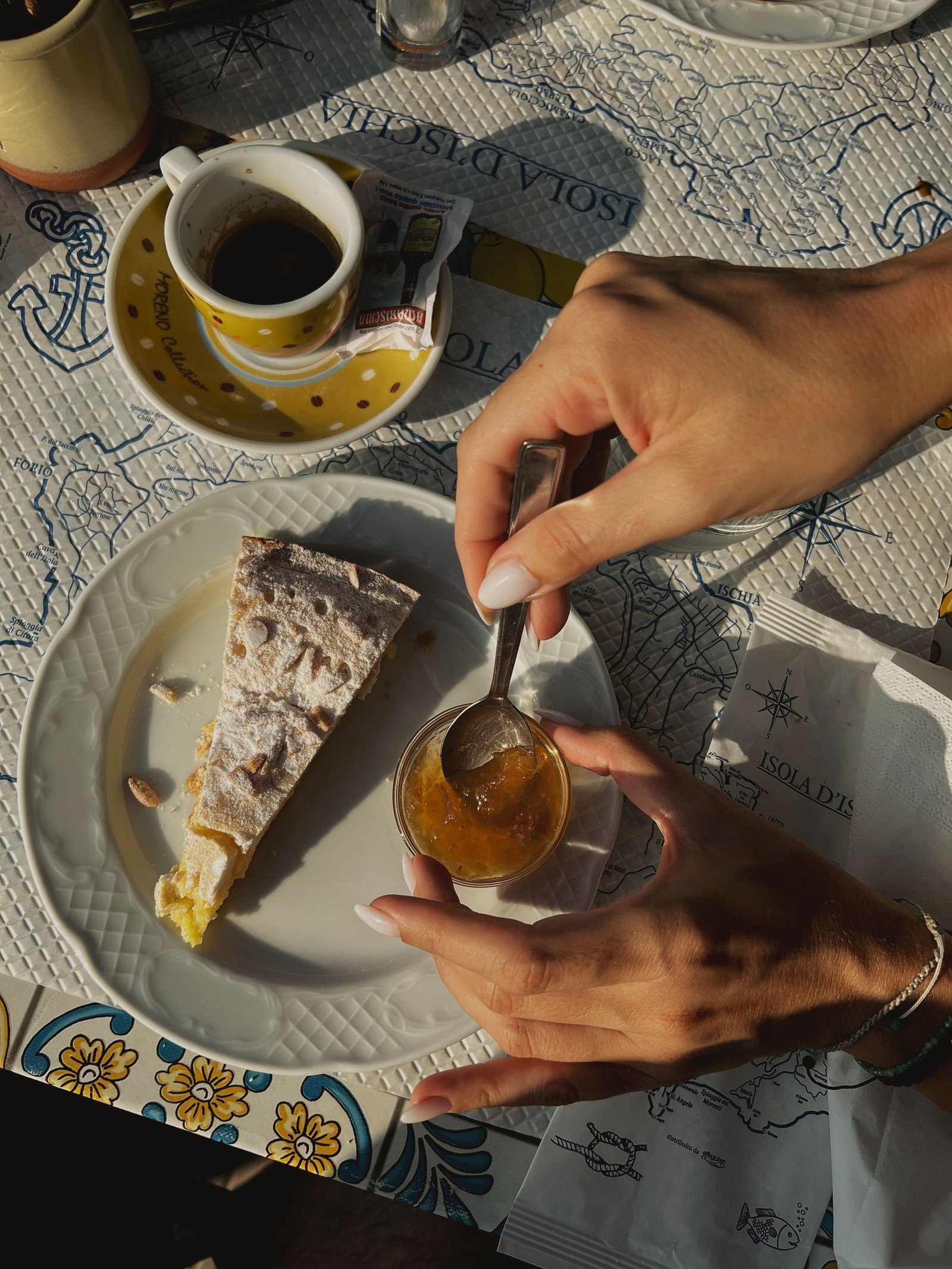 Hands sharing a sunlit dessert beside an espresso on a map of Ischia