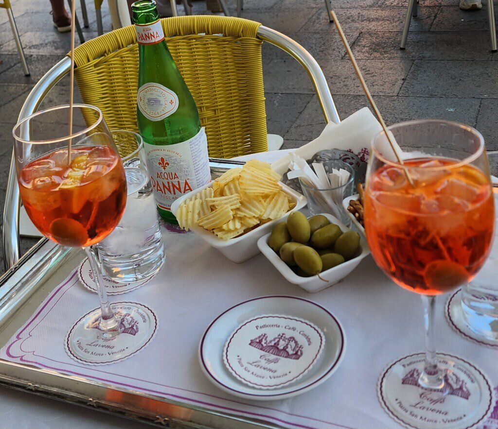 Two orange aperitivo drinks with olives and chips at a Venice café table
