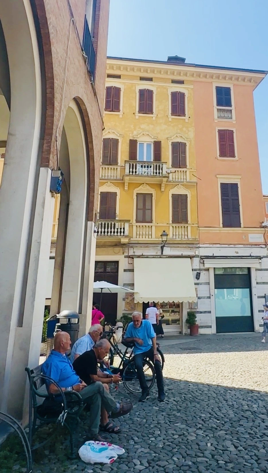 Men chatting beside a bench and bicycles in an Italian piazza beneath yellow and peach buildings