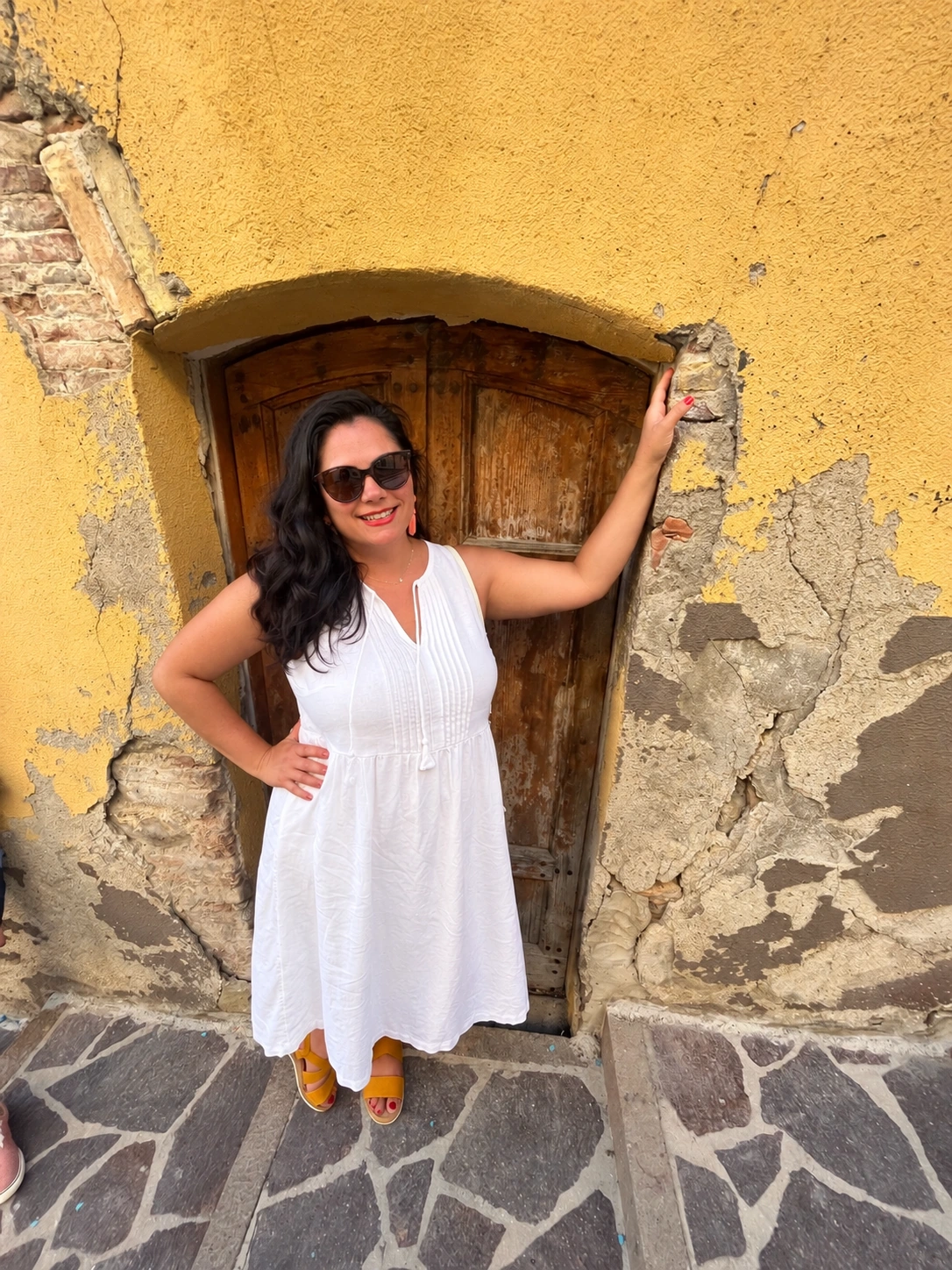 Tiffany in a white dress beside a wooden doorway and sunlit yellow wall in Italy